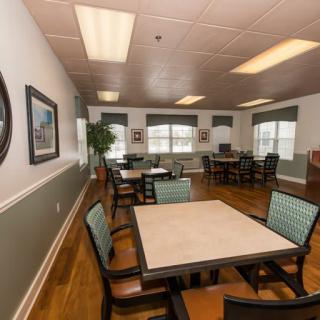 A well-lit dining area with multiple tables and chairs arranged neatly, featuring large windows and a wooden floor.