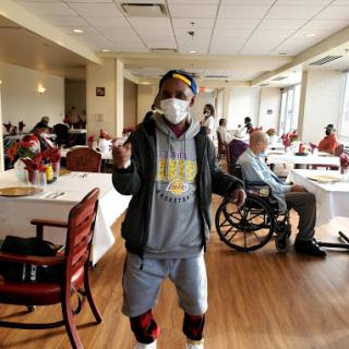 A person in a mask dances in a community dining area while others enjoy their time.