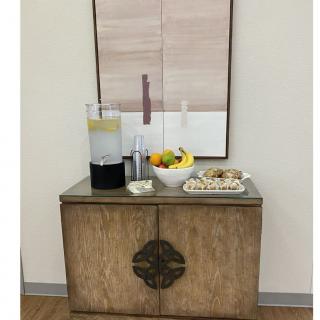 A refreshment station featuring a beverage dispenser, fruit bowl, and cookies on a wooden cabinet with artwork above.