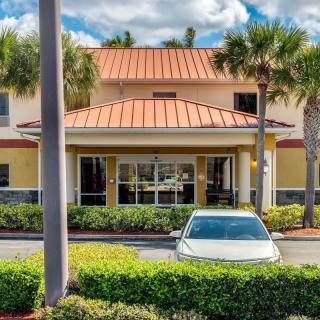 A building entrance with a car parked in front, surrounded by palm trees and landscaping under a partly cloudy sky.