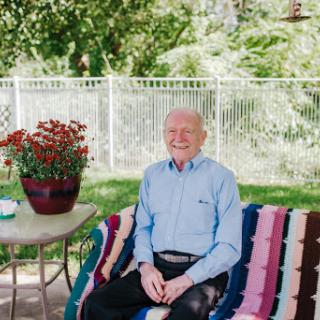 An elderly man enjoying a sunny day outdoors, seated on a colorful blanket with vibrant flowers nearby.