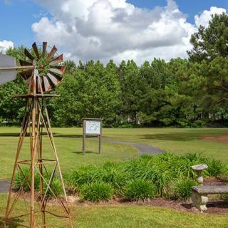 A rustic windmill stands in a lush garden with vibrant greenery and a backdrop of trees under a cloudy sky.