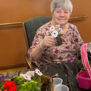 An elderly woman is happily engaging in gardening, holding a small potted plant indoors.