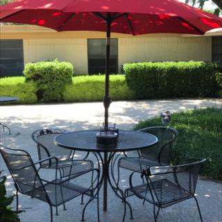A cozy outdoor dining setup featuring a round table with four chairs and a large red umbrella, surrounded by lush greenery.