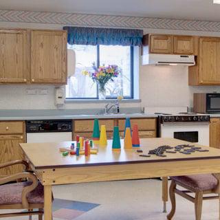 A bright and spacious kitchen featuring a dining table with colorful objects, wooden cabinetry, and a window adorned with flowers.