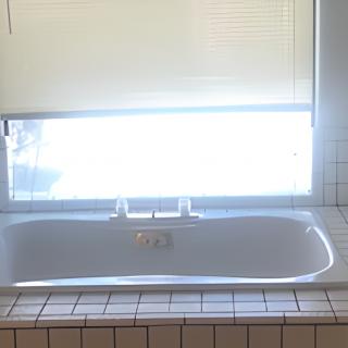 A sunlit bathroom featuring a white bathtub beneath a window with blinds, surrounded by tiled walls.