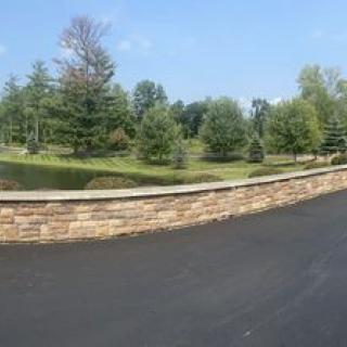 A scenic landscape featuring a stone wall alongside a road, with trees and a pond in the background.