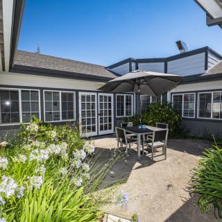 A cozy outdoor patio with a table, chairs, and an umbrella, surrounded by a garden and large windows of the house.