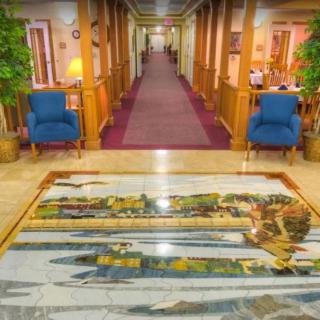 A brightly lit hallway with blue chairs, plants, and a decorative carpet.