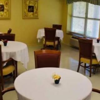 A quiet dining room with round tables and chairs arranged neatly, featuring white tablecloths and a window allowing in natural light.