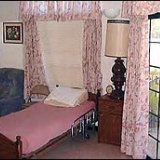 A cozy bedroom features a single bed with a pink cover, a lamp on a wooden nightstand, and floral curtains framing the window.