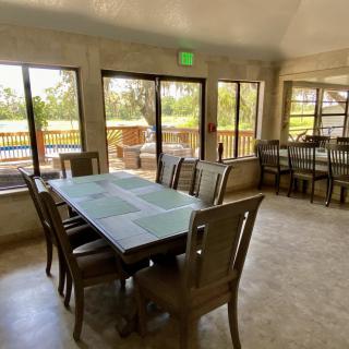 A spacious dining room with a large table and chairs, featuring ample natural light from surrounding windows and a view of the outdoor deck.