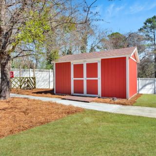 A red garden shed in a backyard with a lawn, tree, and white fence.