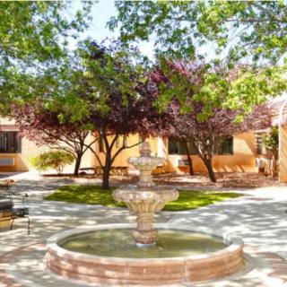 A serene courtyard featuring a central fountain surrounded by trees, benches, and dappled sunlight.