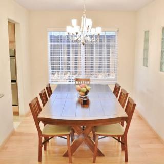 A bright dining room featuring a wooden table with eight chairs, illuminated by a chandelier, and adorned with a fruit bowl.