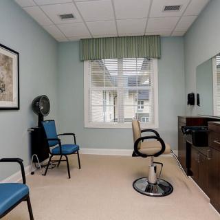 A small salon room with a styling chair, mirror, and hair dryer, featuring a window and waiting area chairs.