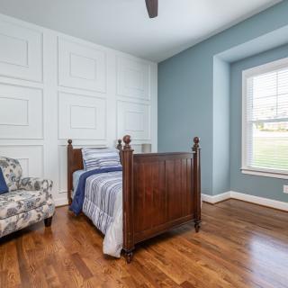 A cozy bedroom with a single wooden bed and an armchair, featuring blue walls and a large window.