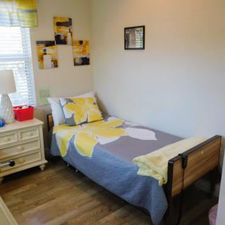 A cozy bedroom with a single bed adorned with yellow and gray bedding, a nightstand with a lamp, and photos on the wall.