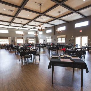An empty dining hall with tables and chairs arranged neatly, featuring a wooden floor and large windows.