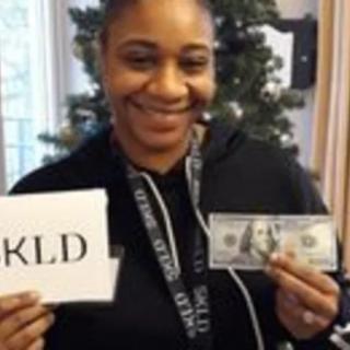 A person smiling while holding a sign and a hundred-dollar bill indoors.