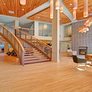 A spacious and modern lobby with wooden flooring, a grand staircase, stylish lighting fixtures, and comfortable seating around a fireplace.