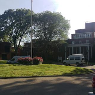A view of a building surrounded by trees and flowers, with vehicles parked nearby.