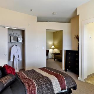A cozy bedroom with a neatly made bed, a dresser, and an open closet revealing a white robe. Soft lighting from a lamp in the adjacent room adds warmth to the space.