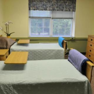 A hospital room with two neatly made beds and small tables, featuring a window with curtains.