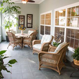 A cozy sunroom featuring wicker furniture and lush green plants, bathed in natural light.