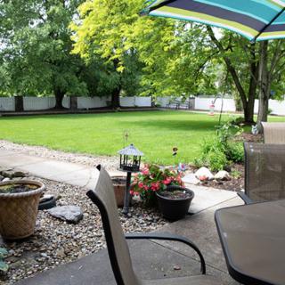 A peaceful backyard patio with chairs, a table under an umbrella, and a well-maintained lawn surrounded by trees.