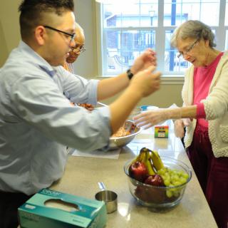 Three people preparing food together in a kitchen with a bowl of fruit on the counter.