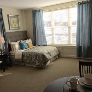A cozy bedroom with a neatly made bed near a large window, adorned with blue curtains and stylish decor.
