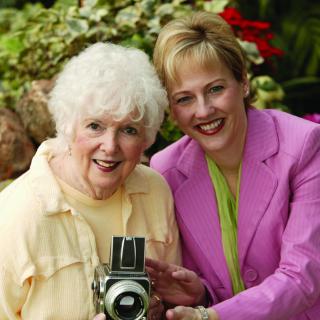 An elderly woman and a younger woman smiling while holding a camera in a garden setting.