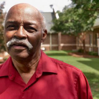 An elderly man wearing a red shirt standing outside in a garden.