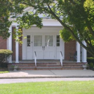 Front entrance of a brick building with steps and columns surrounded by trees and bushes.