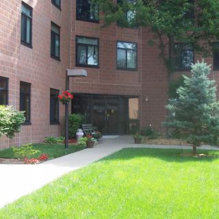 An apartment building with a brick exterior and landscaped entrance surrounded by greenery.
