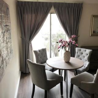 A cozy dining room with a round table, upholstered chairs, and elegant curtains framing a window. A vase with pink flowers adds a decorative touch.