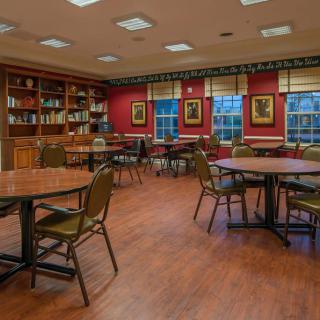 A cozy library meeting room with tables and chairs, surrounded by bookshelves and adorned with paintings.