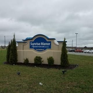 Entrance sign for 'Sunrise Manor', surrounded by landscaped greenery on a cloudy day.
