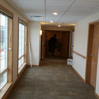 A view of a well-lit hallway with carpet flooring and large windows along one side.