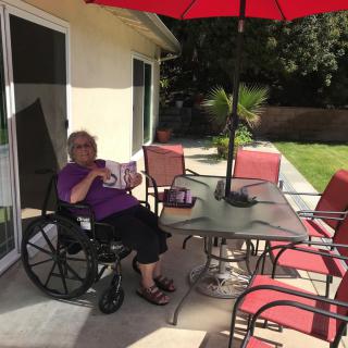 A person in a wheelchair enjoys reading on a sunny patio with red garden furniture and an umbrella.