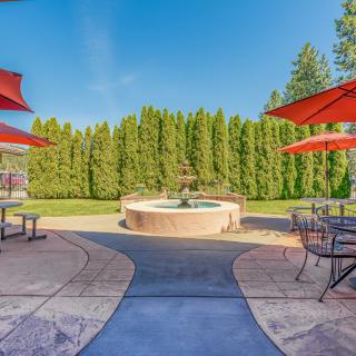 A scenic outdoor patio featuring a central fountain surrounded by tables with red umbrellas and lush greenery.
