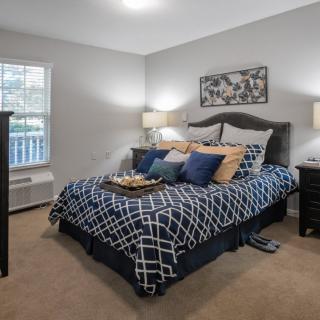 A cozy bedroom featuring a neatly made bed with patterned bedding and pillows, flanked by bedside tables with lamps, a window letting in natural light, and carpeted flooring.