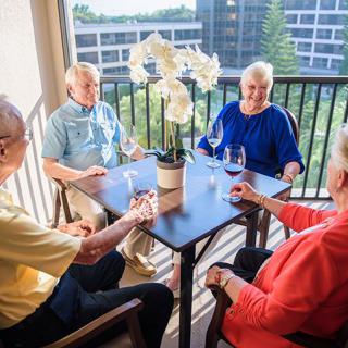 A group of seniors enjoying a conversation on a balcony with wine glasses and a potted orchid on the table.