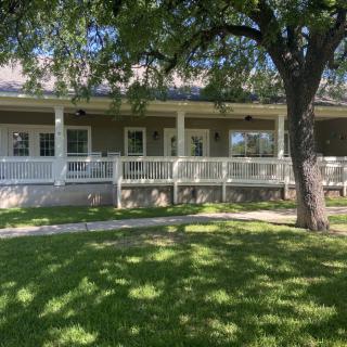 A sunny view of a house with a large porch and tree-shaded lawn.