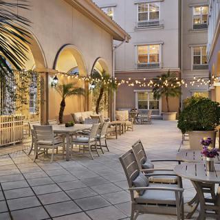 A cozy outdoor patio with string lights, tables, and chairs surrounded by palm trees and building arches.