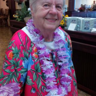 An elderly woman wearing a colorful floral shirt and lei, smiling indoors.