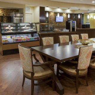 A cozy cafe interior with a large dining table and chairs, featuring a counter with a display case of food and drinks.
