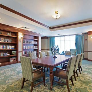 A well-furnished library or conference room featuring a large meeting table surrounded by chairs, with bookshelves lining the walls and a window providing natural light.