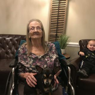 An elderly woman in a wheelchair holding a small dog, with a baby seated nearby in a living room setting.